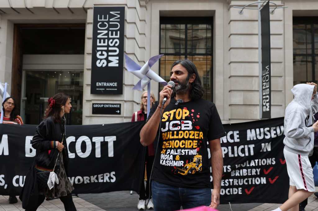 Photograph of speaker wearing a 'Stop JCB' t-shirt, outside the Science Museum entrance, with people holding a large 'boycott' banner in the background.