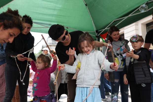 Photograph of children playing with giant bubbles in a gazebo. Smiling adults bend forward to help children holding giant bubble wands. On the left a small child is concentrating hard as they hold the wand aloft. In the middle a child looks down and grins, next to them another holds their hands up to bubbles drifting by.