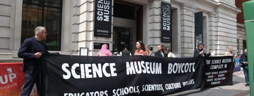 Photograph, taken outside the entrance to the Science Museum where various people are hoding a large black banner with white text in the style of the Science Museum branding, which says: 'Science Museum boycott. Educators, Schools, Scientists, Culture workers.'