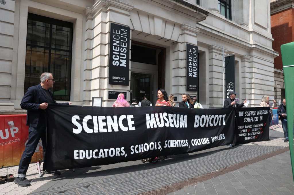 Photograph, taken outside the entrance to the Science Museum where various people are hoding a large black banner with white text in the style of the Science Museum branding, which says: 'Science Museum boycott. Educators, Schools, Scientists, Culture workers.'